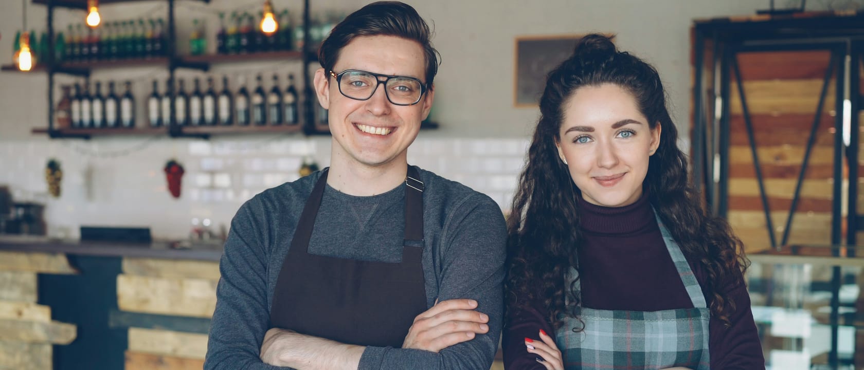 Two smiling owners in aprons stand with arms folded in front of the counter of their small cafe