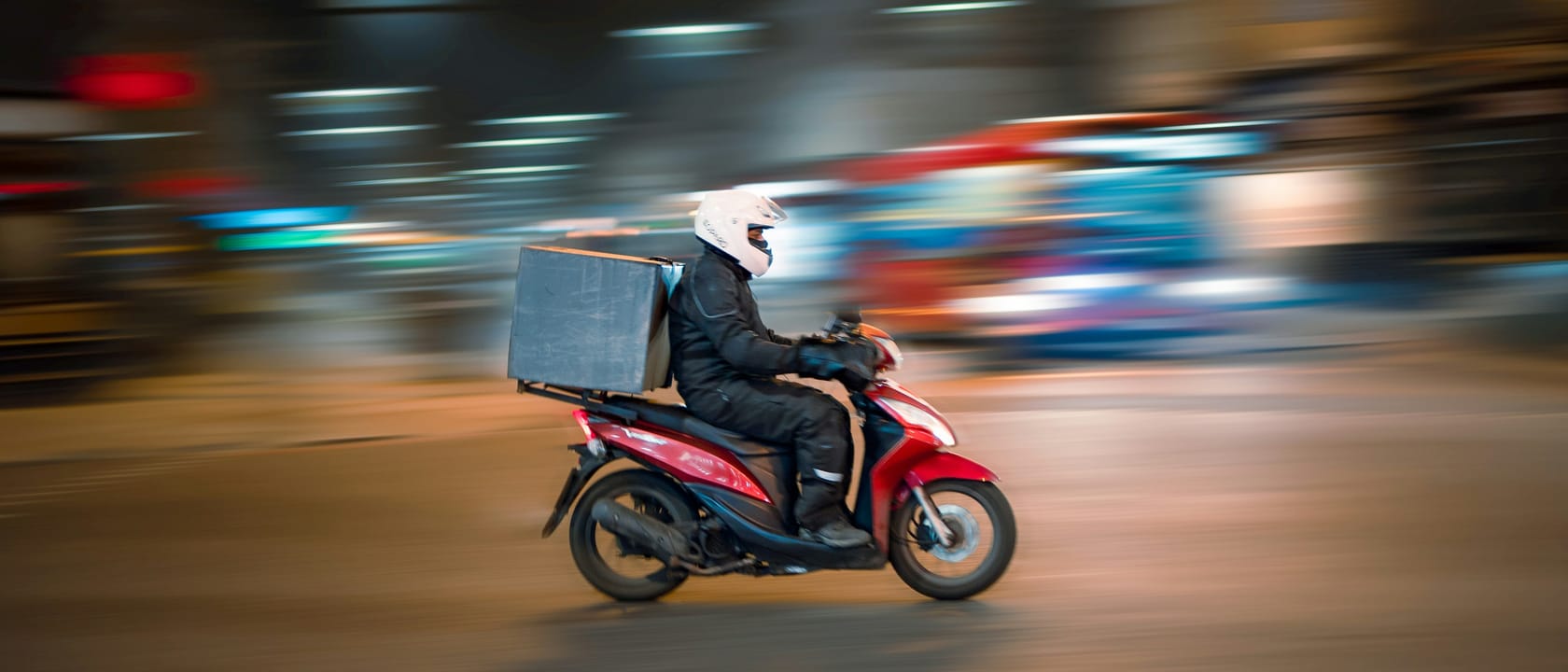 A delivery rider on a red scooter with a plain box on the back, riding through a London street at night with the background blurred by motion