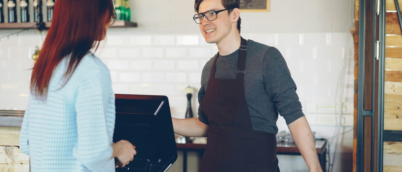 A customer orders at a cafe counter while a smiling member of staff in an apron stands at a touchscreen till